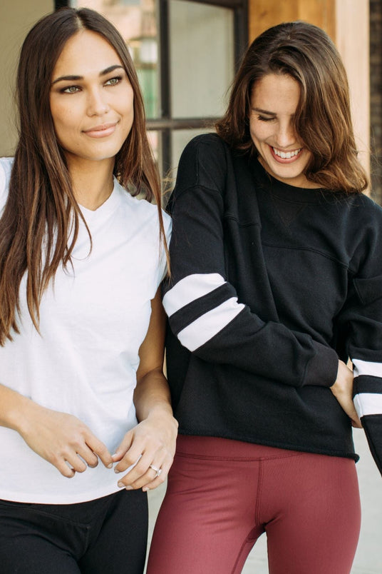Two women standing outdoors, one in a white shirt and black pants, the other in a black sweater with white stripes and red pants.